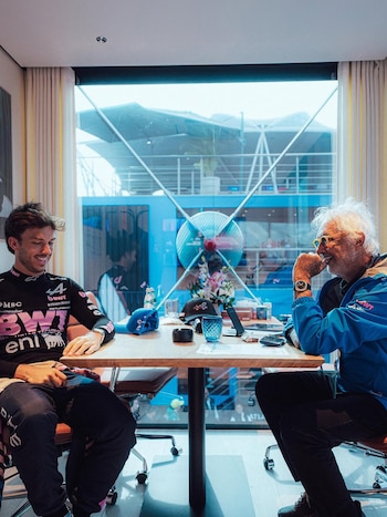 Pierre Gasly y Flavio Briatore conversan sentados frente a frente en una mesa de madera junto a una gran ventana