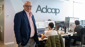 Un hombre con chaqueta azul y anteojos posa junto al letrero de Adcap en una oficina con personas trabajando en computadoras
