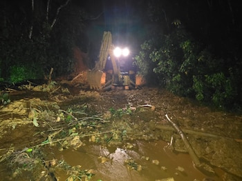 Excavadora amarilla con luces encendidas trabajando en la noche, moviendo lodo, ramas y tierra tras un deslizamiento en una zona boscosa