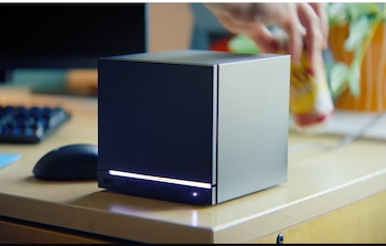 Una Steam Machine cúbica, de colores oscuro y plata, sobre un escritorio con luz frontal y puertos USB. Un ratón negro y una mano desenfocada están al fondo