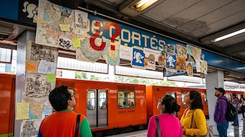 Un letrero en el Metro Villa de Cortés, cubierto por un collage con mapas, ilustraciones históricas y signos de interrogación rojos, es observado por pasajeros mientras un tren naranja espera.