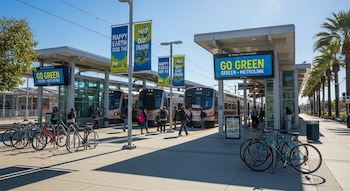 Estación de tren moderna en el sur de California con tres trenes Metrolink detenidos, pasajeros, bicicletas estacionadas y carteles de "Happy Earth Day".