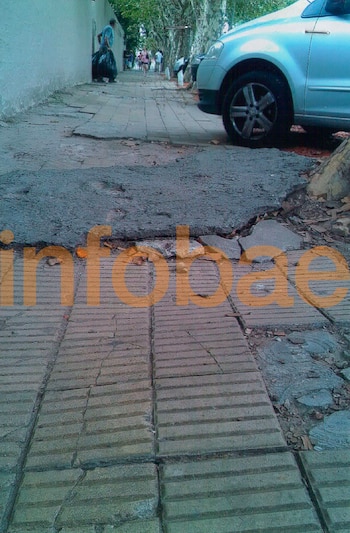 Vereda deteriorada con baldosas rotas y tierra, un tronco de árbol en el lateral y la rueda de un automóvil, con el logo de Infobae