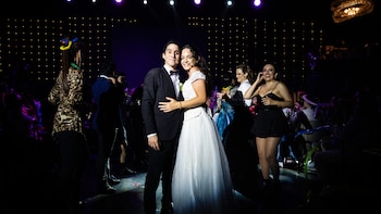Un hombre con traje oscuro y una mujer con vestido de novia blanco sonríen abrazados en un escenario iluminado, rodeados por varias personas