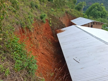 Vista aérea de una empinada ladera de tierra rojiza erosionada cubierta de vegetación, junto a dos techos de metal corrugado gris claro de estructuras