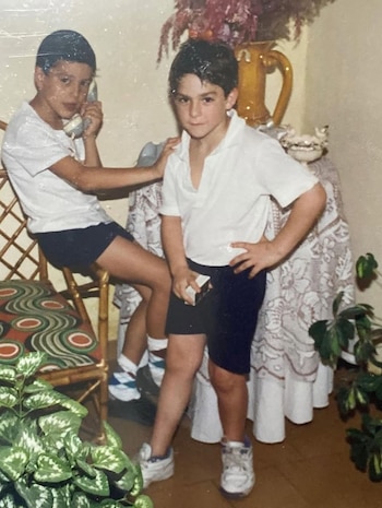 Dos niños, uno sentado en silla de madera con teléfono y otro de pie con las manos en la cintura y un objeto. Fondo con mesa y plantas