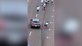 Vista aérea de un SUV gris detenido en una carretera, con una persona en el suelo a su lado. Otros individuos caminan más adelante en la vía