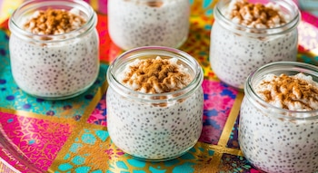 Cinco vasitos de cristal con arroz con leche y chía, cubiertos con canela en polvo, dispuestos sobre una bandeja multicolor con patrones dorados, rosados y turquesas.