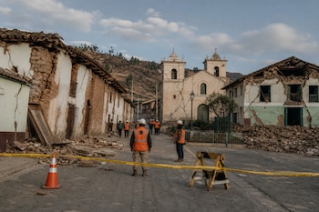 Calle empedrada con casas agrietadas, escombros, una iglesia al fondo, cinta de seguridad amarilla y personas con chalecos naranjas.