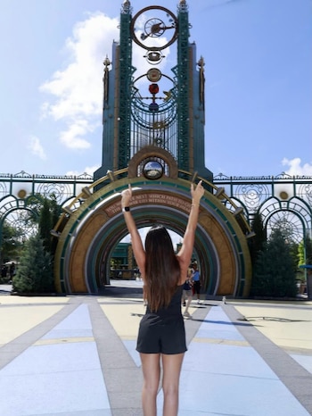 Antonela Roccuzzo de espaldas, con un short y top negros, levanta los brazos frente a una majestuosa entrada arqueada con elementos decorativos y un reloj