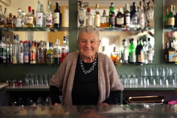 Retrato de una mujer de edad avanzada sonriendo detrás de una barra con botellas y vasos en los estantes
