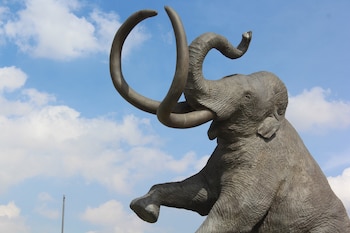 Escultura de mamut con grandes colmillos curvos y trompa levantada, vista desde abajo contra un cielo azul claro con nubes blancas