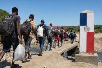 Personas con mochilas caminan en fila sobre un puente junto a un hito fronterizo pintado con la bandera de República Dominicana.