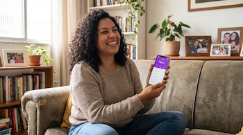 Mujer de cabello rizado oscuro y tez morena sonríe ampliamente, sentada en un sofá, usando la aplicación Nubank en su teléfono. Hay libros y plantas en el fondo.
