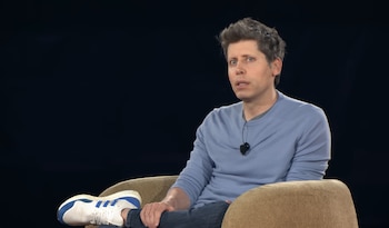 Sam Altman sentado en una silla, vestido con camiseta azul claro, pantalones vaqueros y zapatillas blancas. Tiene un micrófono prendido en la camisa