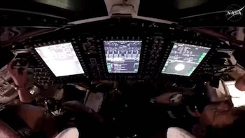 Vista interior de una cabina espacial oscura con múltiples pantallas brillantes que muestran datos técnicos y un cuerpo celeste; manos de un astronauta operando controles