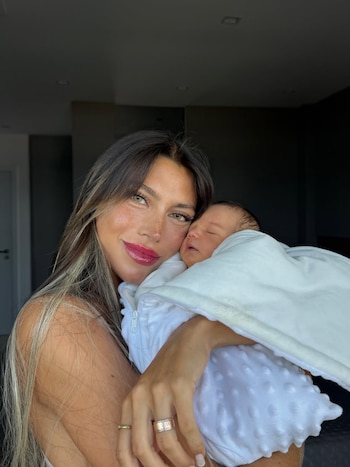 Una mujer sonríe a la cámara mientras sostiene a un bebé dormido envuelto en una manta blanca