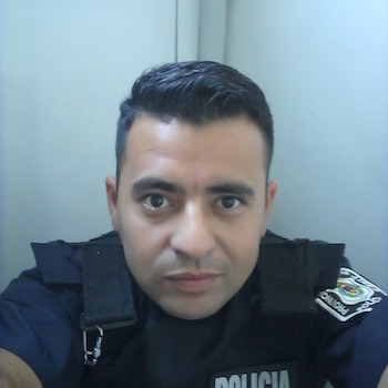 Primer plano de un hombre con uniforme de policía y chaleco antibalas oscuro, mirando directamente a la cámara. Tiene cabello oscuro corto