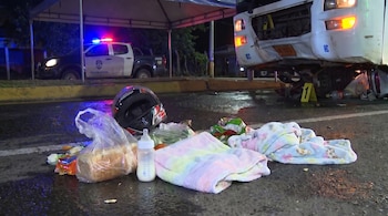 Imagen nocturna de una escena de accidente con un camión blanco dañado, una patrulla policial con luces encendidas, un casco, una pacha y dos mantas de bebé sobre el asfalto mojado.