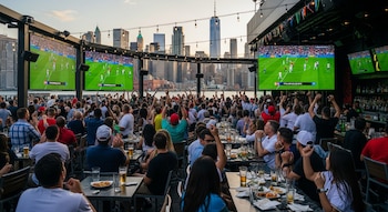 Gran multitud de personas viendo un partido de fútbol en pantallas gigantes en un bar al aire libre con la silueta de Nueva York de fondo al atardecer.