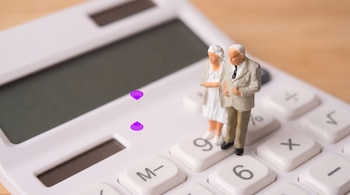 Una calculadora blanca con pantalla oscura y dos figuras en miniatura de un hombre y una mujer ancianos. Dos puntos púrpuras flotan sobre las teclas.
