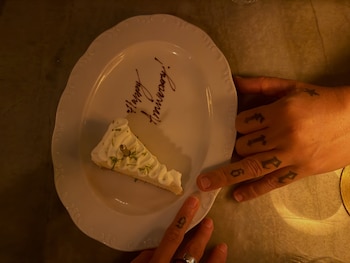 Un plato blanco con una porción de pastel, el mensaje Happy Anniversary escrito en chocolate y dos manos con tatuajes que sostienen el borde