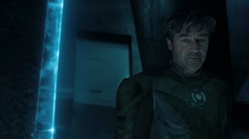 Primer plano de un hombre con traje oscuro con un símbolo circular en el pecho, junto a una columna vertical de luz azul brillante en un espacio oscuro