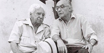 Fotografía en blanco y negro de dos hombres mayores sentados. Uno, con cabello blanco y camisa clara, sostiene gafas y un sombrero. El otro usa camisa a rayas y gafas