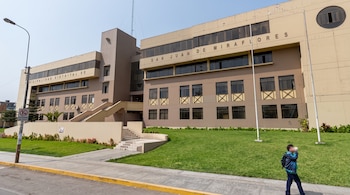 Edificio beige con el texto Municipalidad Distrital de San Juan de Miraflores, jardín frontal, rampas y un transeúnte en la vereda.
