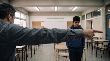 Un hombre extiende el brazo e indica la salida a un joven con la cabeza agachada en un aula con pupitres y sillas