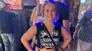 Mckenna Kindred, una mujer rubia sonriente, vestida con un top negro estampado, posa frente a un fondo borroso con otras personas en un local