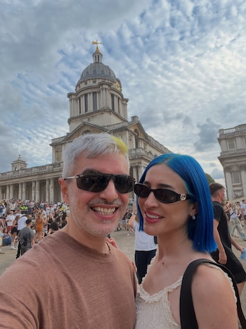 Un hombre y una mujer con cabello azul y anteojos de sol sonríen frente a un gran edificio antiguo con cúpula y una multitud detrás
