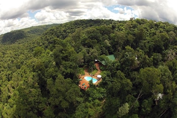Vista aérea de un complejo con edificios de techos verdes, una piscina azul con tumbonas y caminos serpenteantes, todo inmerso en una vasta selva tropical bajo un cielo nublado
