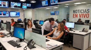 Interior de una sala de redacción de noticias con varias personas trabajando en escritorios. Se ven monitores, teclados, teléfonos y grandes pantallas de televisión mostrando informativos.
