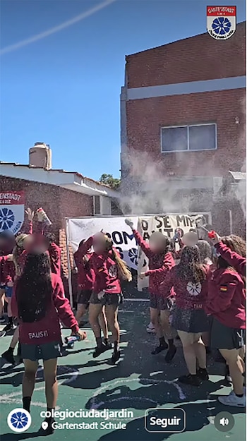 Grupo de estudiantes con uniformes color borgoña en un patio escolar, algunos rociando espuma blanca. Al fondo, un edificio de ladrillo bajo un cielo azul claro