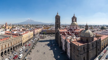 Vista panorámica aérea del centro histórico de Puebla, con la Catedral, el Zócalo, edificios coloniales, tráfico y personas, y el Popocatépetl al fondo.