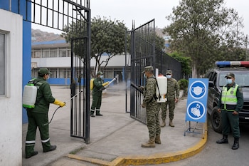 Militares y trabajadores con mascarillas e implementos de fumigación trabajan en la entrada de una escuela con rejas de metal.