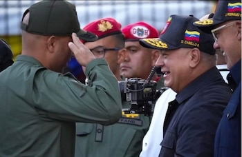 Un militar con gorra verde saluda. Un hombre con gorra negra, bandera de Venezuela y 'D. CABELLO R.' ríe. Militares con boinas rojas y gorras. Una cámara