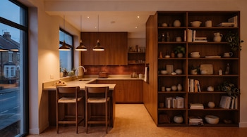 Cocina moderna con mobiliario de madera, encimeras claras, dos taburetes de bar, fregadero, lámparas colgantes y una estantería empotrada con objetos. Se ve una ventana grande.