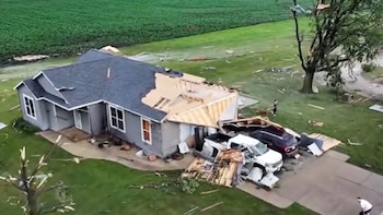 Fotografía aérea de una casa con tejado dañado y madera expuesta, junto a dos coches destrozados en el camino de entrada y un árbol con ramas rotas