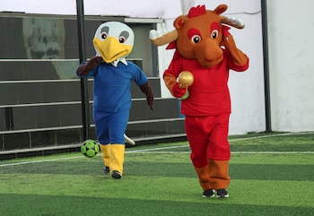Dos personas con disfraces de mascotas, un águila blanca y un toro marrón, caminan sobre césped artificial. El toro sostiene un trofeo dorado y el águila tiene un balón de fútbol verde