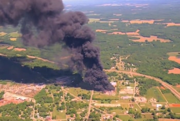 Vista aérea de una gran columna de humo negro elevándose sobre una zona industrial y rural con árboles verdes, campos marrones y casas pequeñas