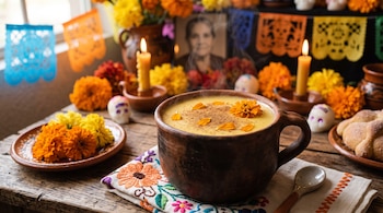 Taza de barro con bebida caliente y pétalos de cempasúchil sobre una mesa decorada con velas, flores, papel picado y pan de muerto.