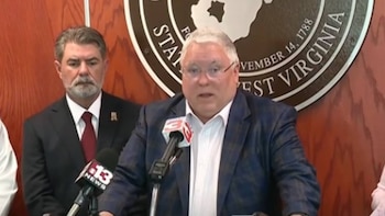 Un hombre con cabello blanco y gafas habla en un podio con micrófonos, un hombre barbudo está detrás y el sello de Virginia Occidental es visible