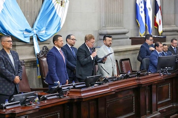 Un hombre en traje oscuro habla en un podio con micrófono y papeles, rodeado de otros hombres y banderas de Guatemala y Honduras al fondo
