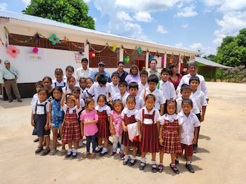 Grupo de niños y adultos de pie frente a una escuela blanca decorada con globos y banderines coloridos bajo un cielo azul con nubes