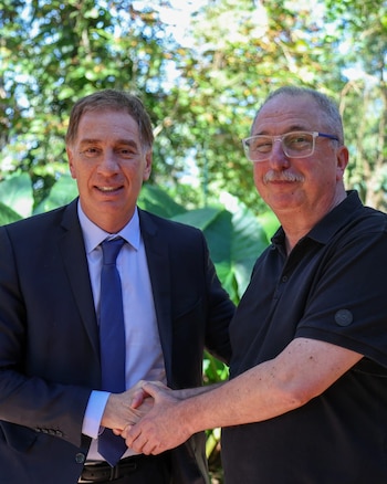 Hugo Passalacqua, en traje oscuro, y Diego Santilli, con polo negro y gafas, se dan la mano sonriendo frente a un fondo verde de árboles