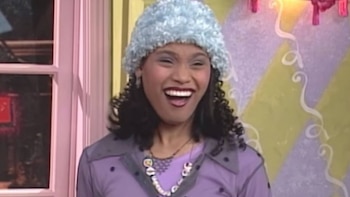 Mujer sonriendo con gorro azul de textura rizada, blusa lila, collar con dijes, y pelo oscuro y rizado; al fondo hay una ventana y una pared bicolor