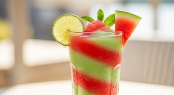 Un vaso de cristal con granizado de sandía rojo y granizado de lima verde en capas diagonales, decorado con una rodaja de lima, un trozo de sandía y hojas de menta.