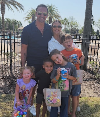 Ryan Lochte y Kayla Rae Reid posan con cinco niños frente a una valla negra, palmeras y una piscina; la niña más pequeña lleva una cesta de huevos de Pascua